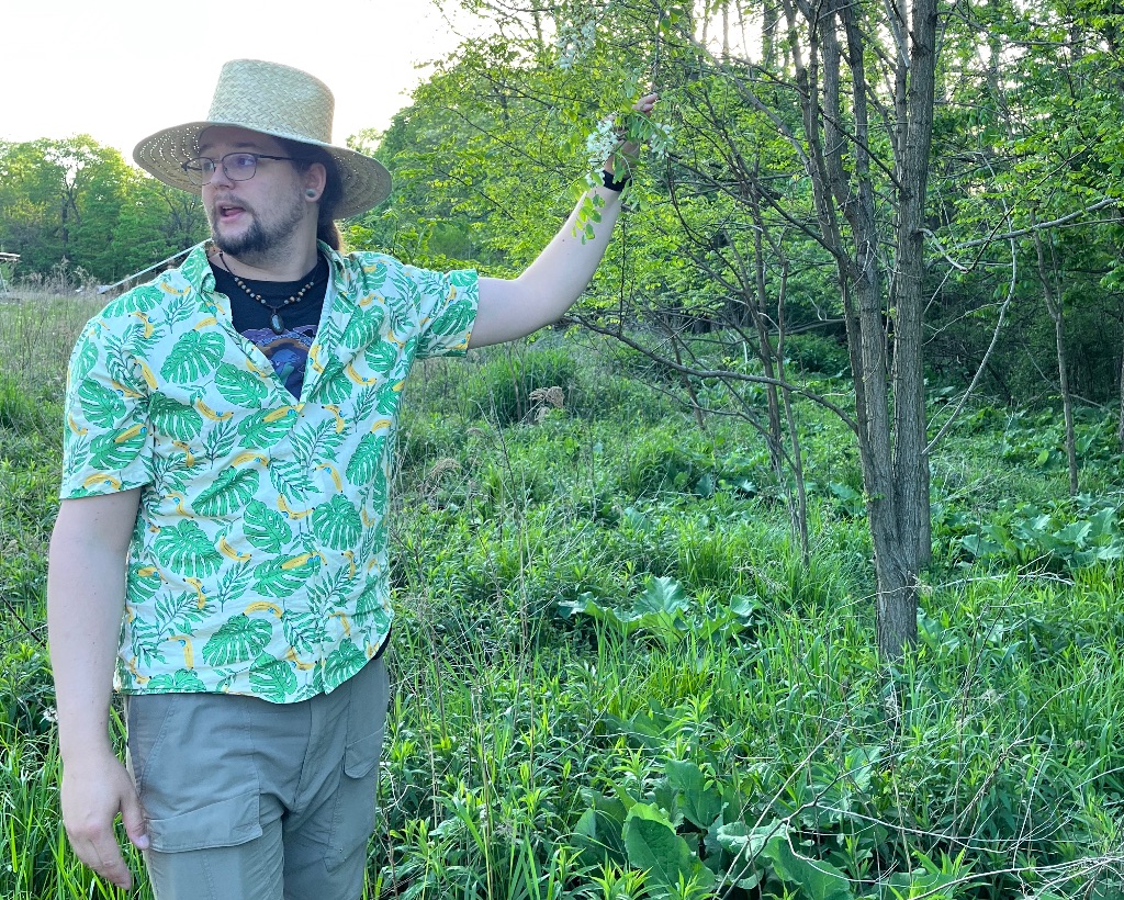 a white man with long brown hair and beard stands in front of a small tree. He wears a patterned green and yellow shirt and a tan brimmed hat. He is holding the leaves of a small tree.
