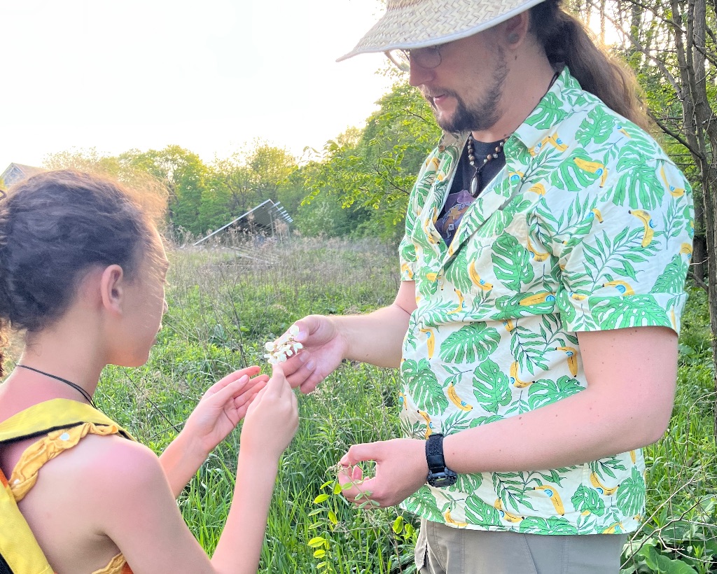 a white man with long brown hair and beard. He wears a patterned green and yellow shirt and a tan brimmed hat. He is holding handing a cluster of small white flowers to a little girl with brown hair and a yellow shirt and backpack.