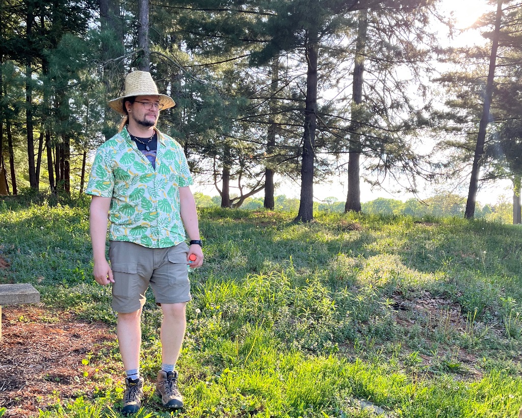 a white man with long brown hair and beard stands on a wooded path with the sun shining behind him. He wears a patterned green and yellow shirt and a tan brimmed hat. There are tall pine trees behind him.