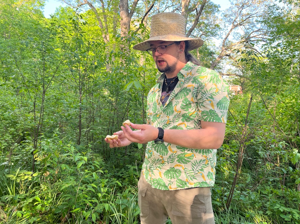 a white man with long brown hair and beard stands on a wooded path. He wears a patterned green and yellow shirt and a tan brimmed hat. He is holding a mushroom.