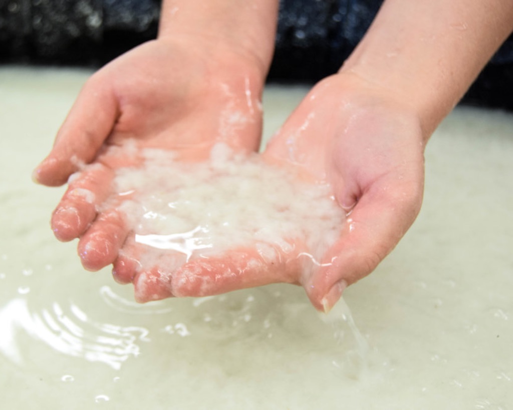 Two white hands hold wet pulp in their hands. Water drips through their fingers into the cloudy water below. 