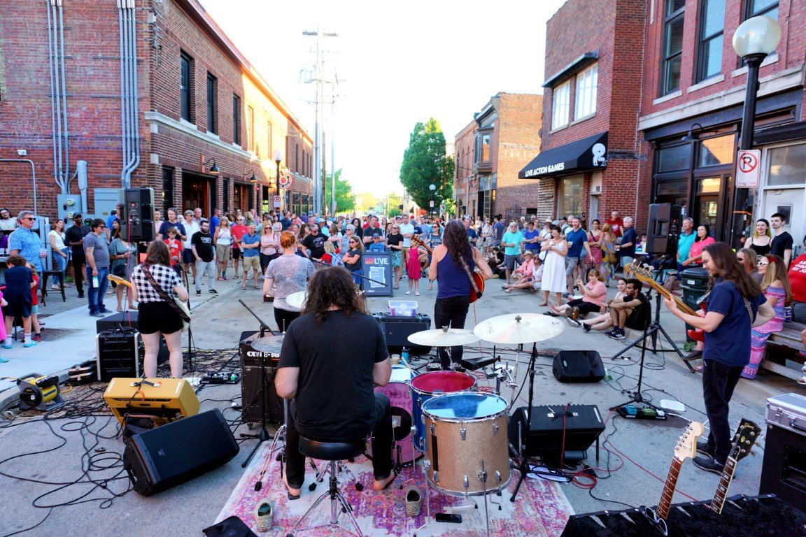 A band is set up in the center of a street, looking out to a crowd of people gathered in front of them. There are brick buildings lining either side of the street.