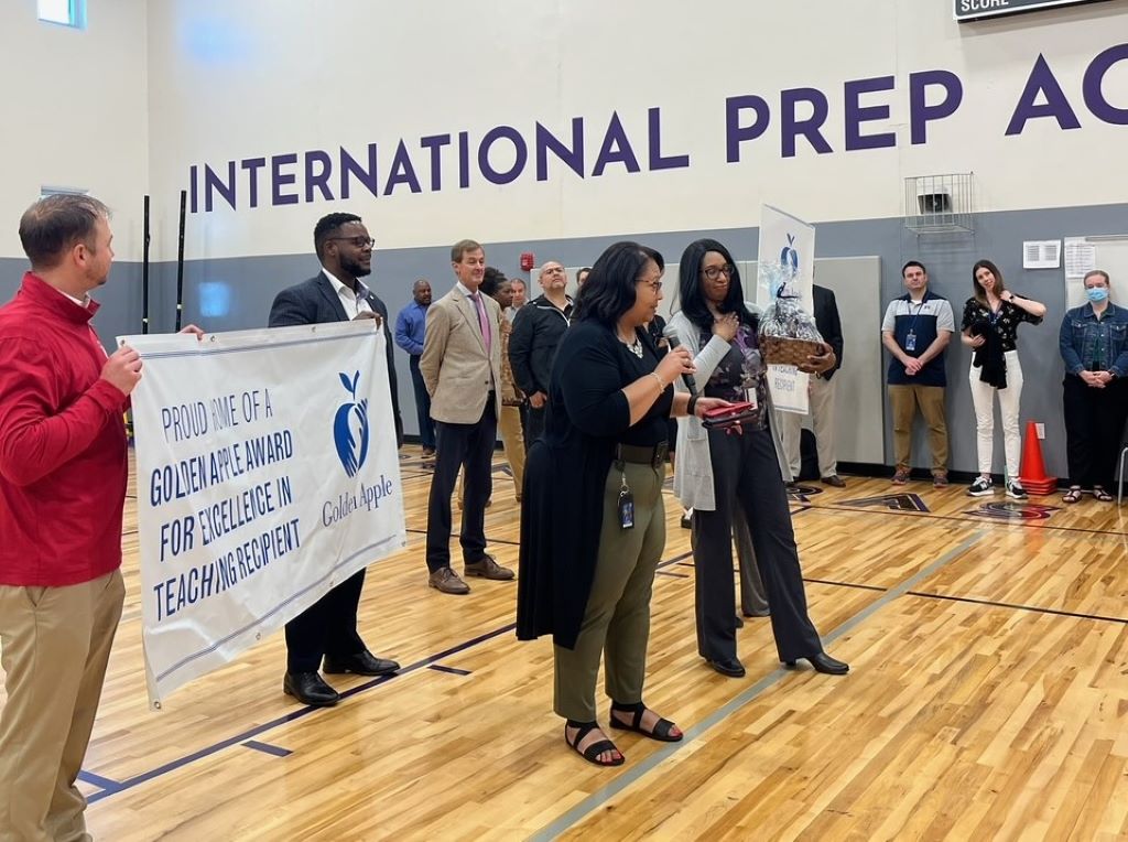 A group of people are gathered on a gym floor. A Black woman is holding a microphone and gesturing towards another Black woman holding an award. There are two men holding a white banner behind them.