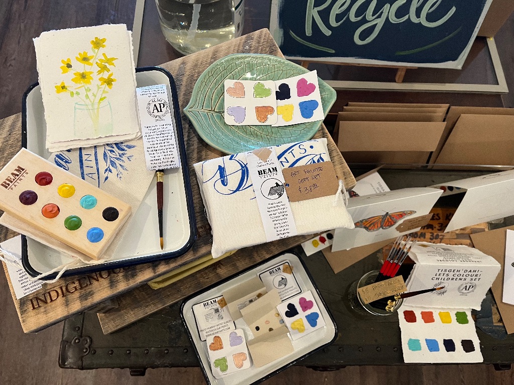 A table is displayed with a variety of paints. Some are heart shaped while others are in round wooden trays. There are also paintbrushes, paper, and a metal tin
