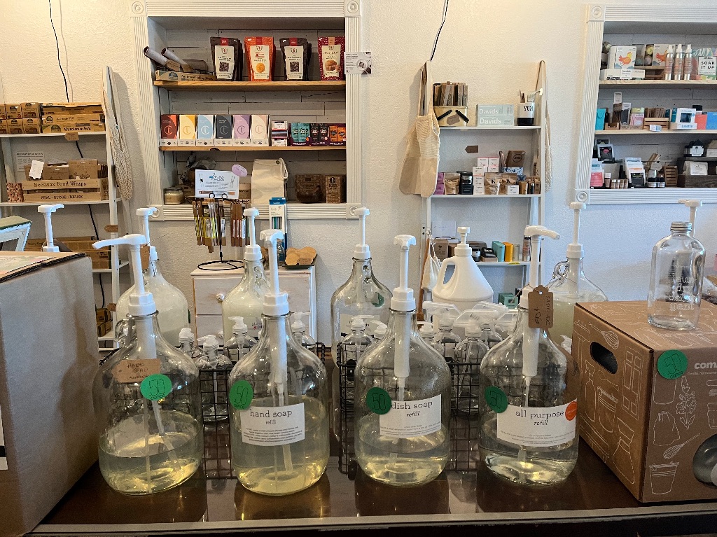 8 large glass bottles full of clear liquids sit on a large table. The bottles have pumps attached and are labeled dish soap, hand soap, and all purpose