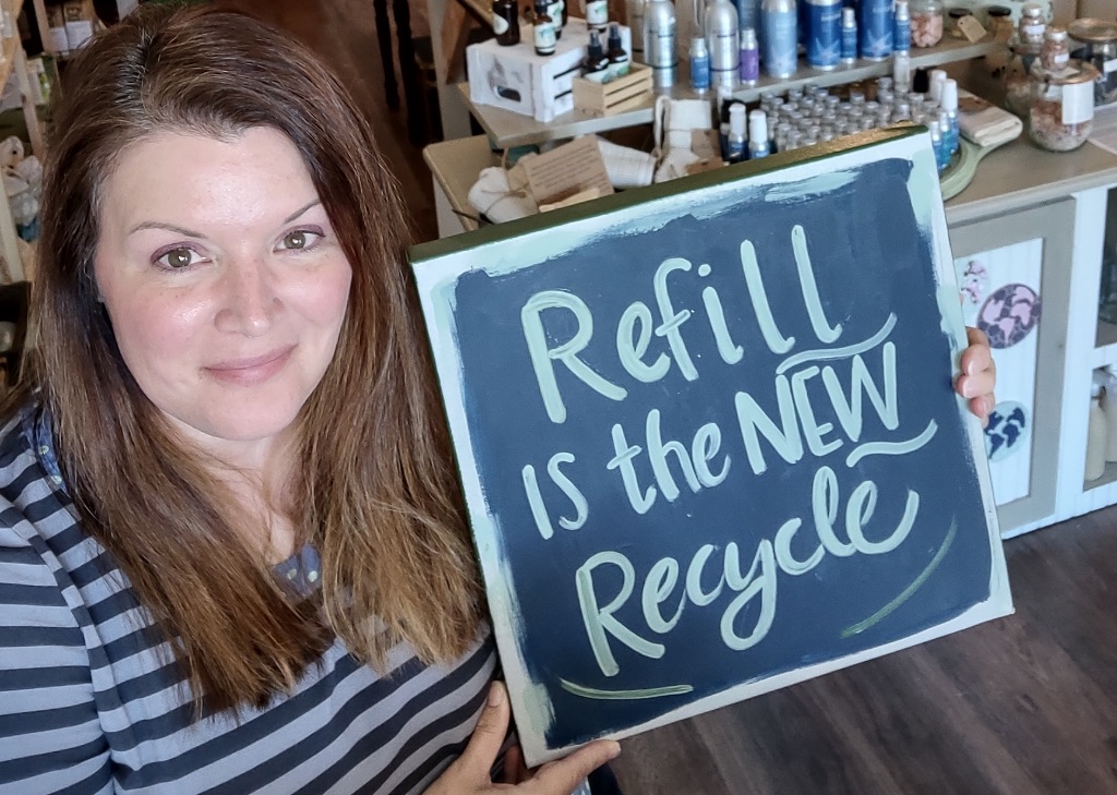 a white woman with long brown hair holds a sign that says, refill is the new recycle she is wearing a grey and black striped shirt.