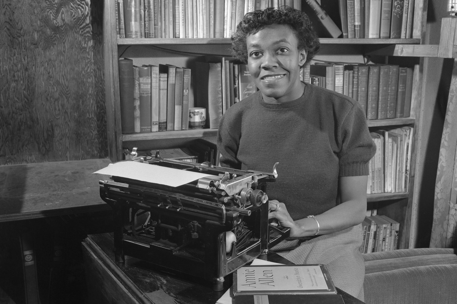 A black and white photograph of a Black woman with short hair, seated behind a typewriter. There is a bookshelf behind her.
