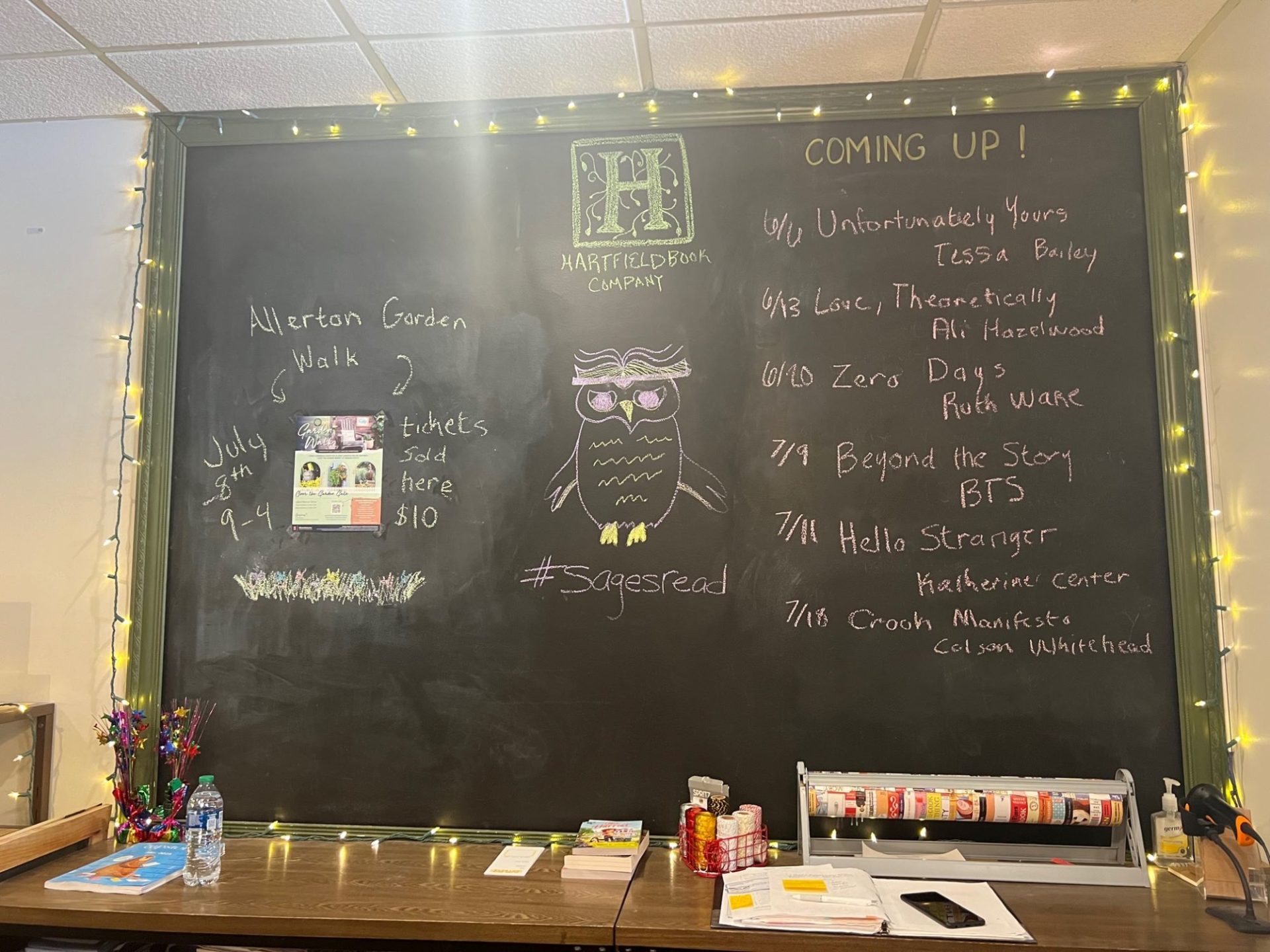 A large chalkboard hangs on a wall, and is lined with yellow string lights. There are several dates and book names written in white chalk.