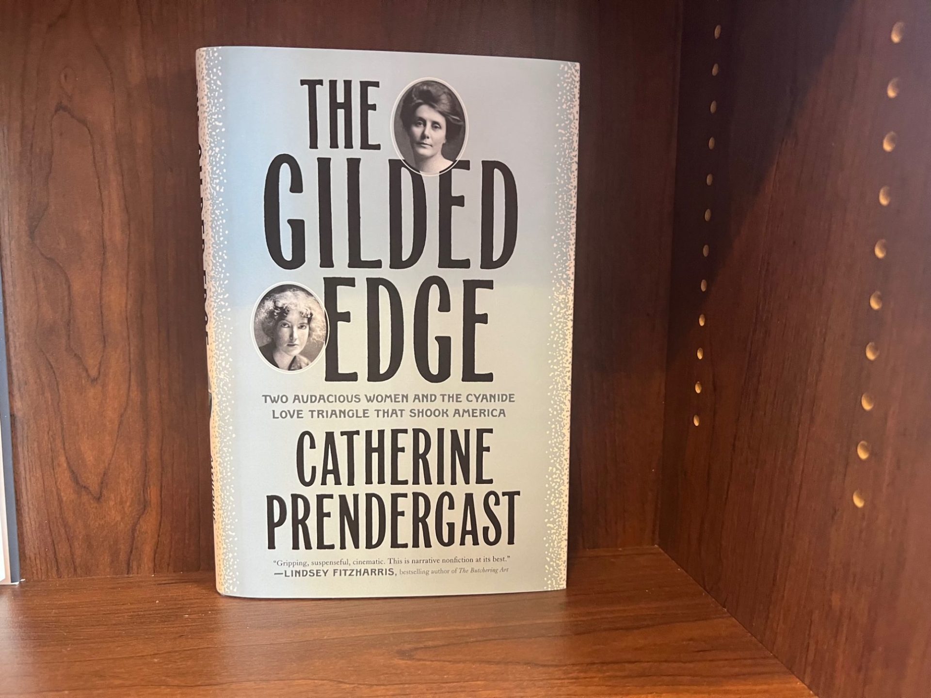 A book with a light gray cover that says The Gilded Edge in black lettering sits on a brown wooden shelf.