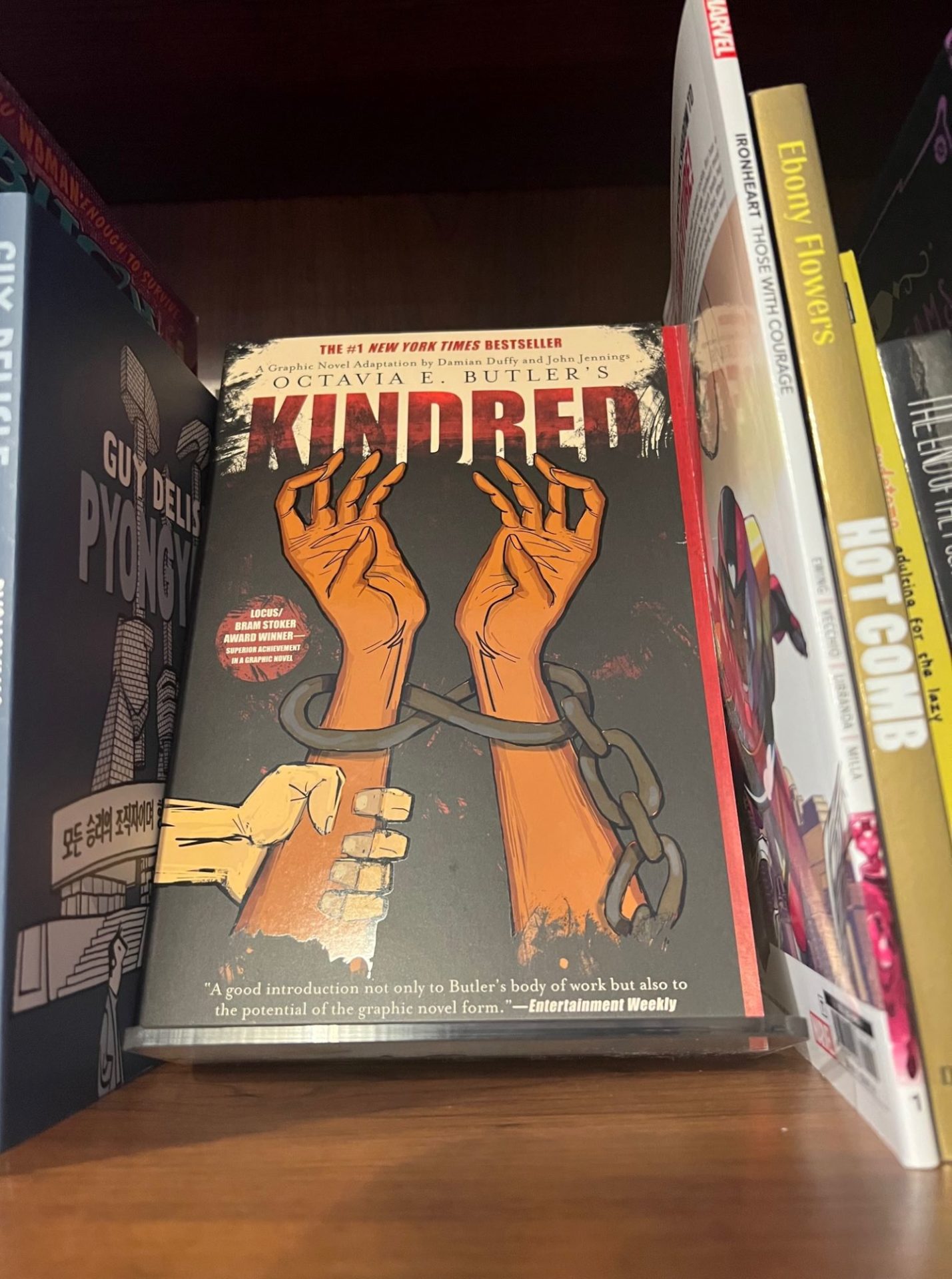 A book with two brown hands bound by handcuffs and the word Kindred in red letters sits on a brown wooden shelf with the spines of other books along either side.