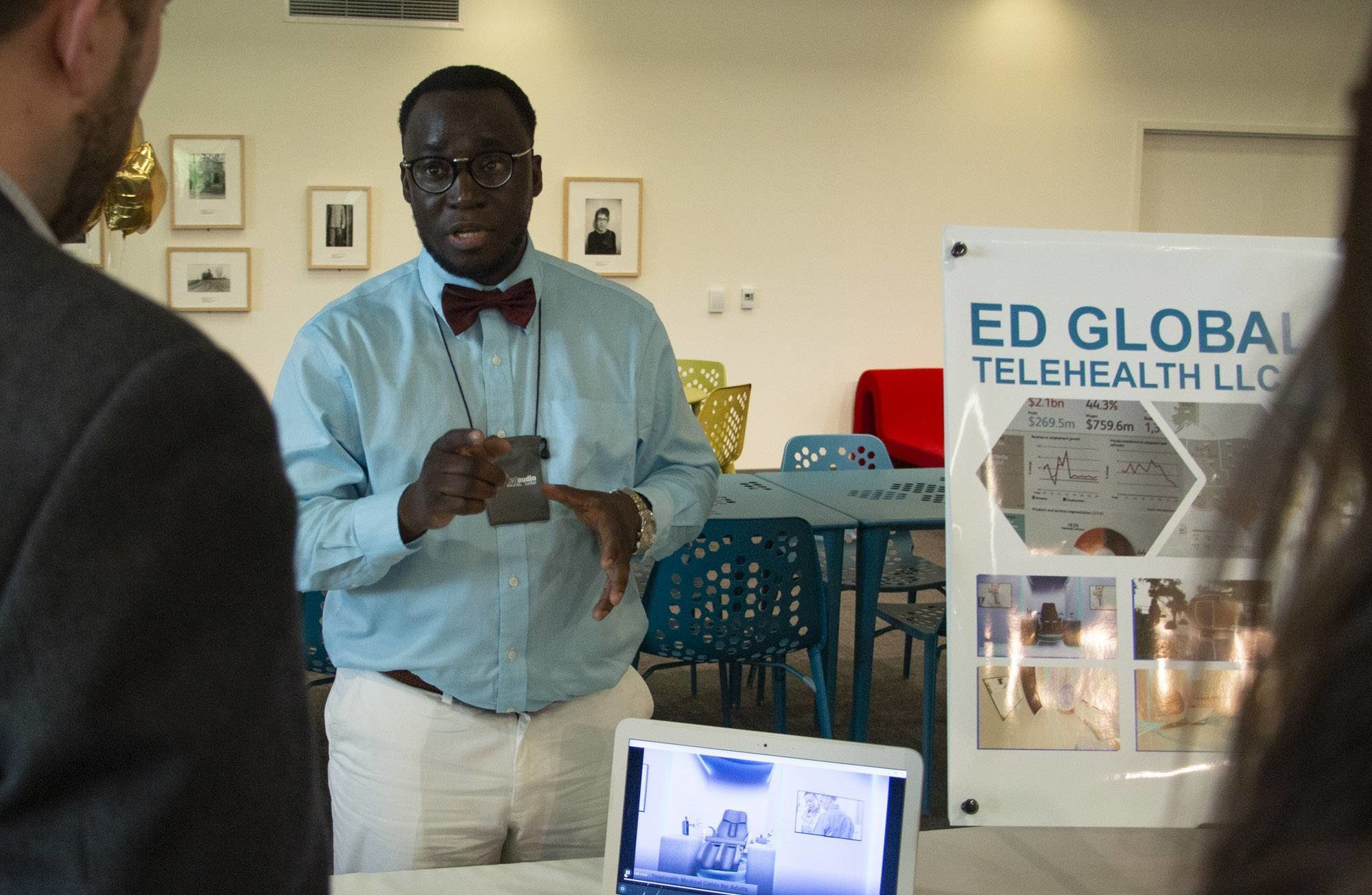 A Black man in a blue shirt and black bowtie is standing behind a table, gesturing and talking to someone.