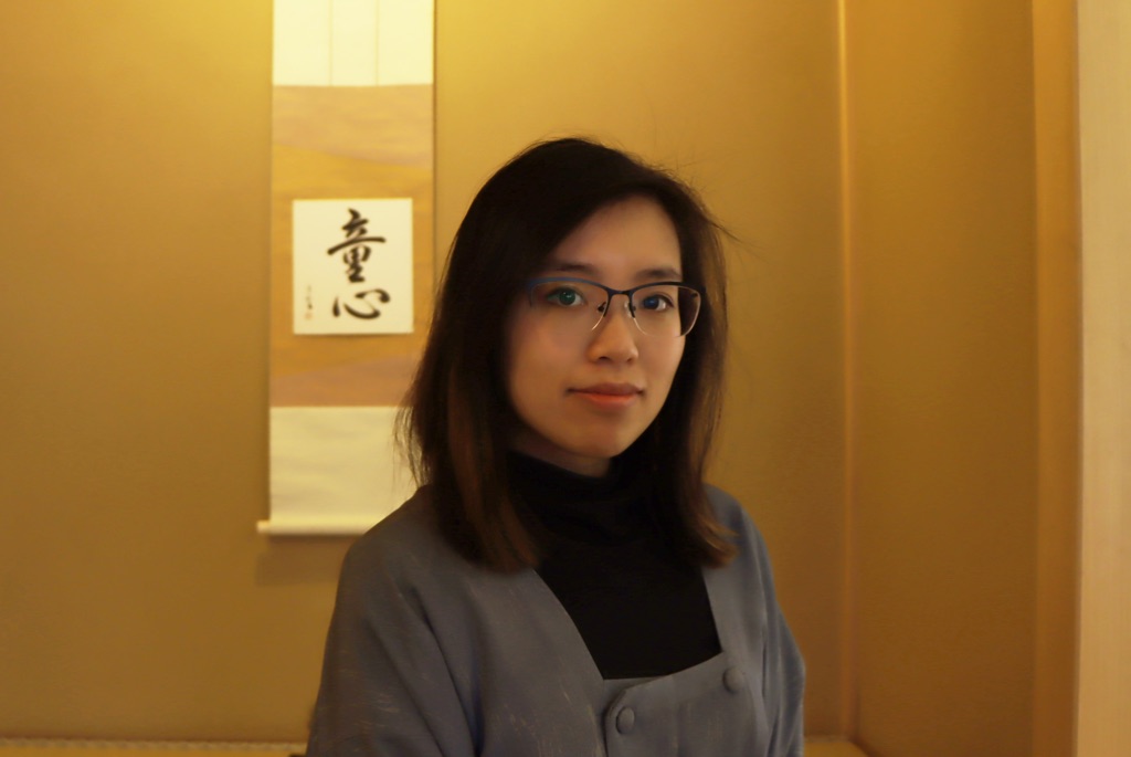 a portrait of a young woman with black framed glasses and dark shoulder length hair stares at the camera. She is wearing a black turtleneck with a gray jacket over it. The walls behind her are brown and have a small square of paper with Japanese calligraphy on it.  