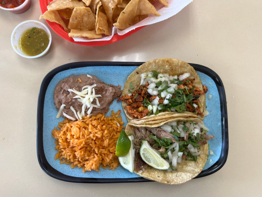 Two tacos are on a ceramic light blue plate with a side of rice and beans. In the top left, there is a red basket of tortilla chips with a plastic cup of salsa at La Mixteca restaurant in Urbana. Photo by Alyssa Buckley