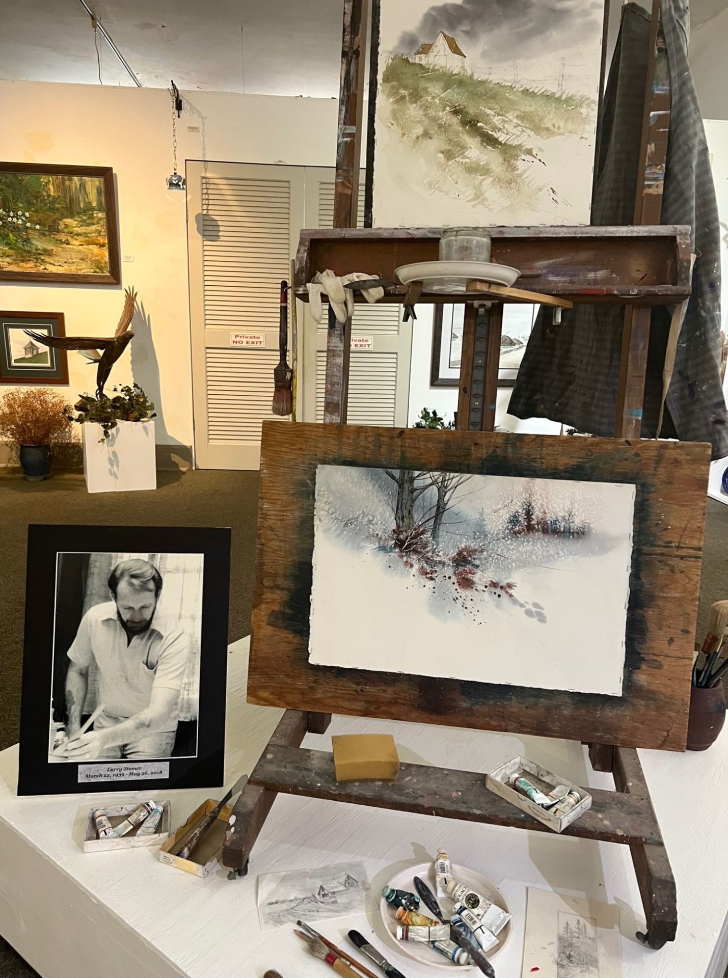 A display with a wooden easel with two paintings propped on it. There are tubes of paint and brushes scattered below it. A black and white photo of a man painting sits alongside it.