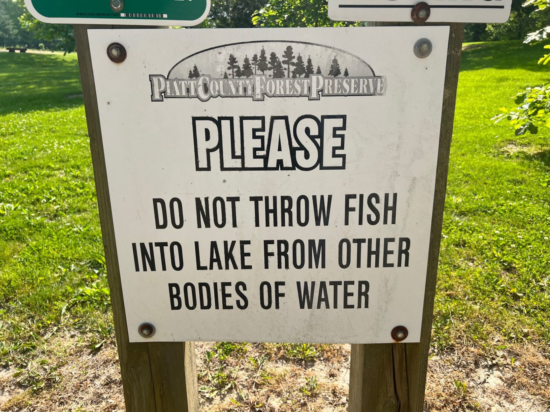 A white sign on wooden posts with black lettering that says Please Do Not Throw Fish Into the Lake From Other Bodies of Water.