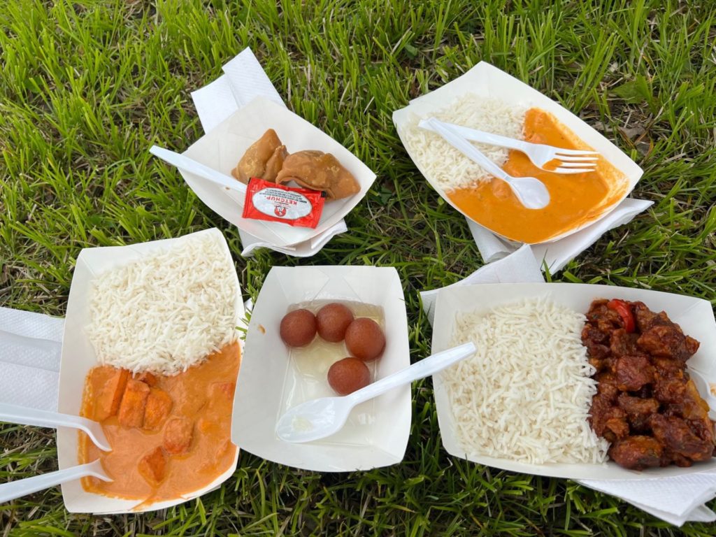 A collection of white cardboard trays hold different Indian foods. Three of them have white rice alongside an entree with sauce. There is one with four round balls of dough, and one with two samosa. 
