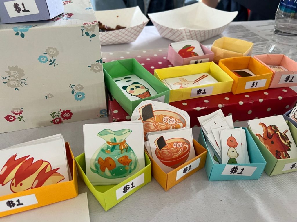 Small colorful paper boxes are sitting on a table. Each box has a stack of stickers, and each has a white label on the front that says $1 in black ink.