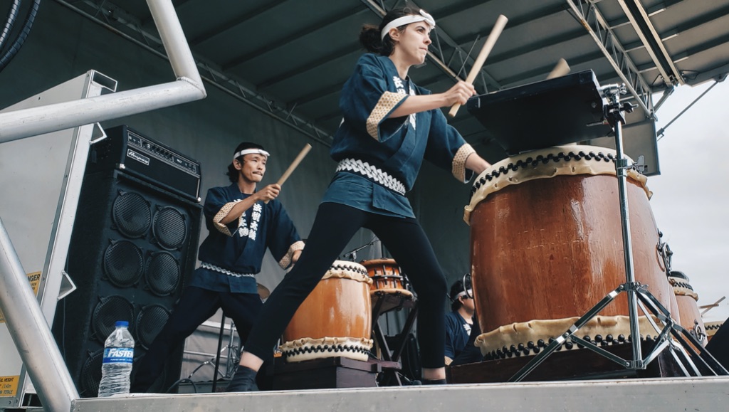 a side view of two performers standing on a stage playing large brown drums with large wooden sticks. They are wearing black pants and dark blue kimonos with white belts and headbands 