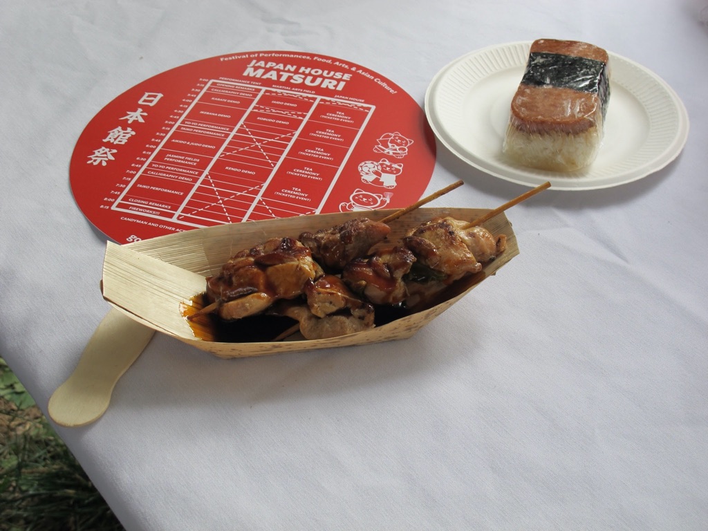 a bamboo dish full of glazed chicken skewers. To the left is a round fan that says "Japan House Matsuri" on the right is a pice of sushi atop white rice and wrapped in nori. 