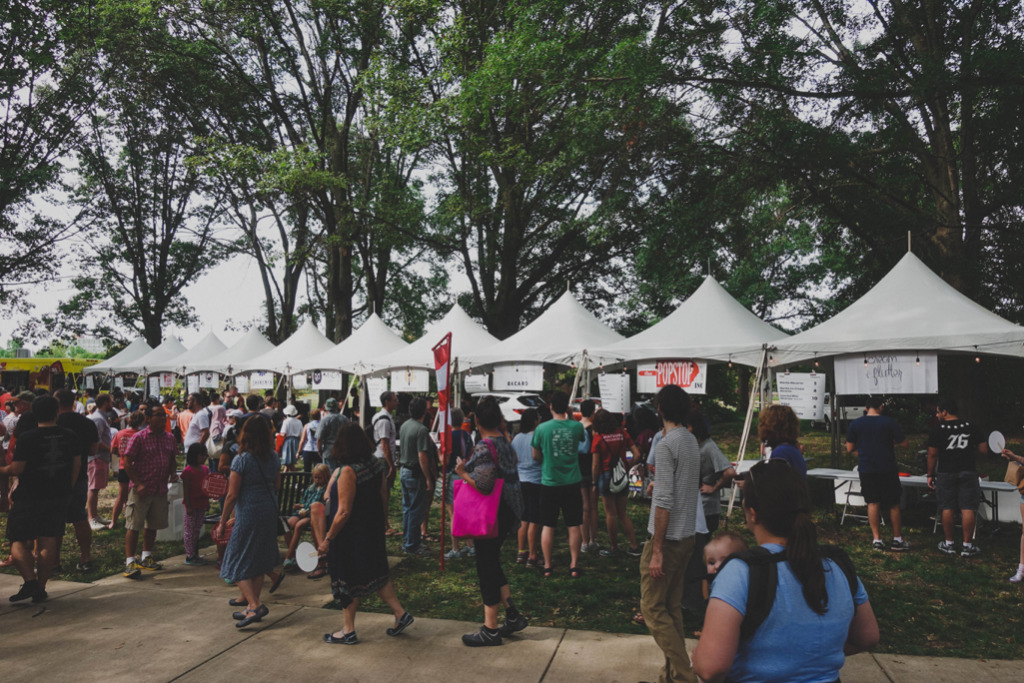 a large group of people walk on the sidewalk past a row of ten white vendor tents. Behind the tents are tall trees with green leaves. 