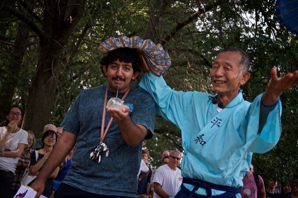 two men stand in the center of the image. the man on the right is tan with gray and clack hair. He is wearing a sky blue kimono tied with a black belt. He has his arms raised to his sides and appears to be laughing. The man on the right has brown skin, dark hair, and a mustache. He is wearing a gray t-shirt with a. lanyard of keys around his neck. He is blowing into a small straw connected to a bubble of candy. 