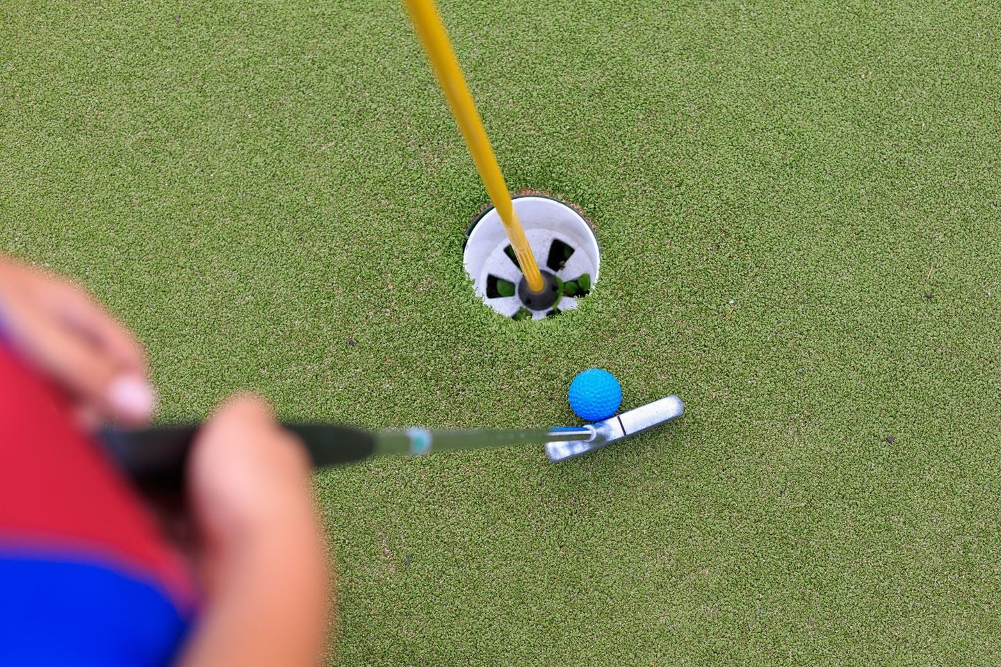A close up of a hole on a putting green, with a yellow flag pole sticking out of it. A person is holding a golf club alongside a bright blue golf ball. 