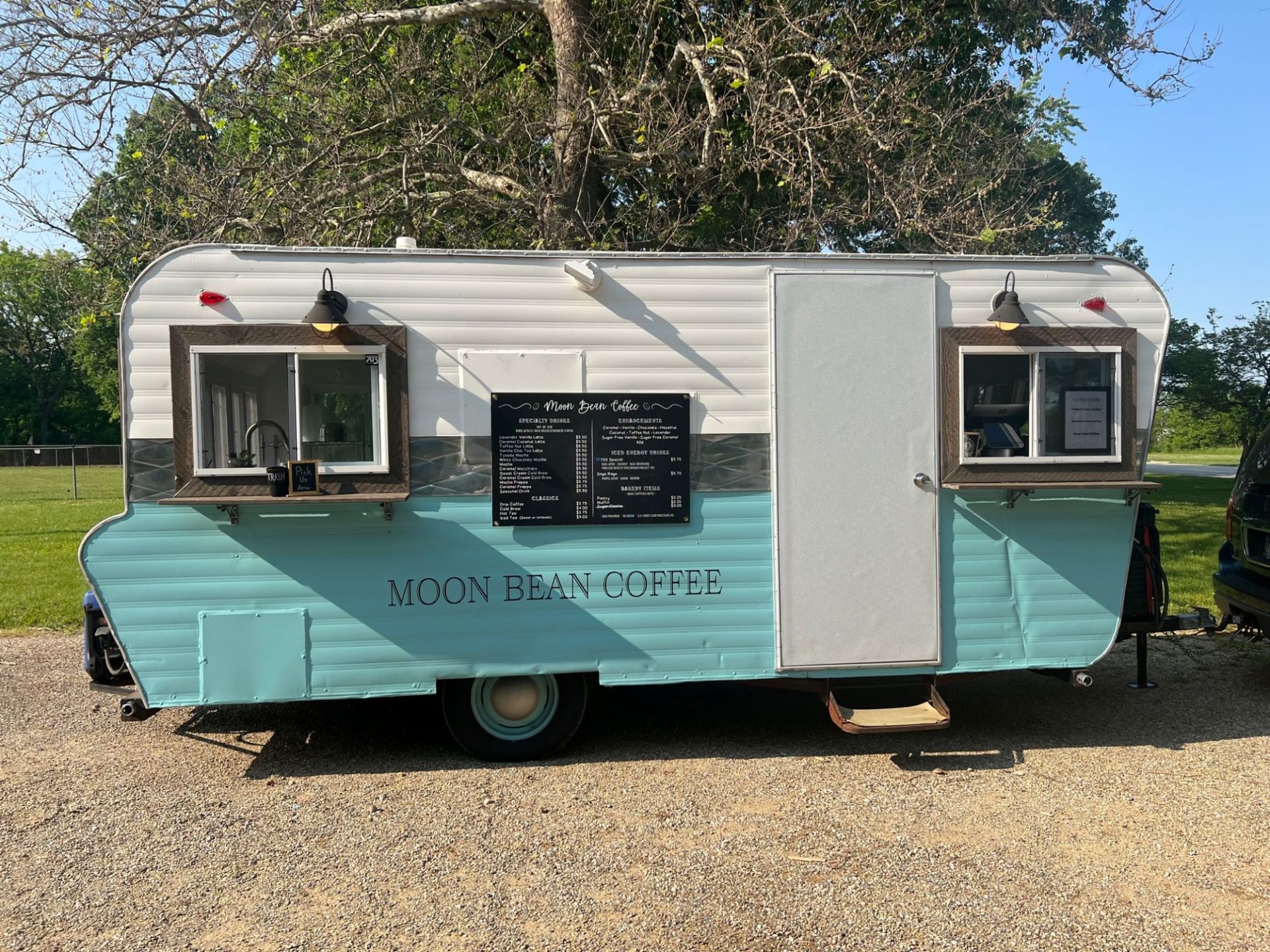 A camping style trailer that is painted white on the top and turquoise on the bottom. It says Moon Bean Coffee in black lettering. There is a door and two windows on the side, as well as a black menu board.