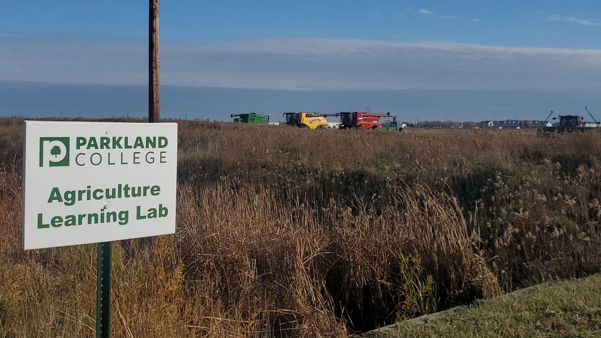 A large farm field with farm machinery in the background. There is a sign in the foreground that is white and says Parkland College Agriculture Learning Lab in green.