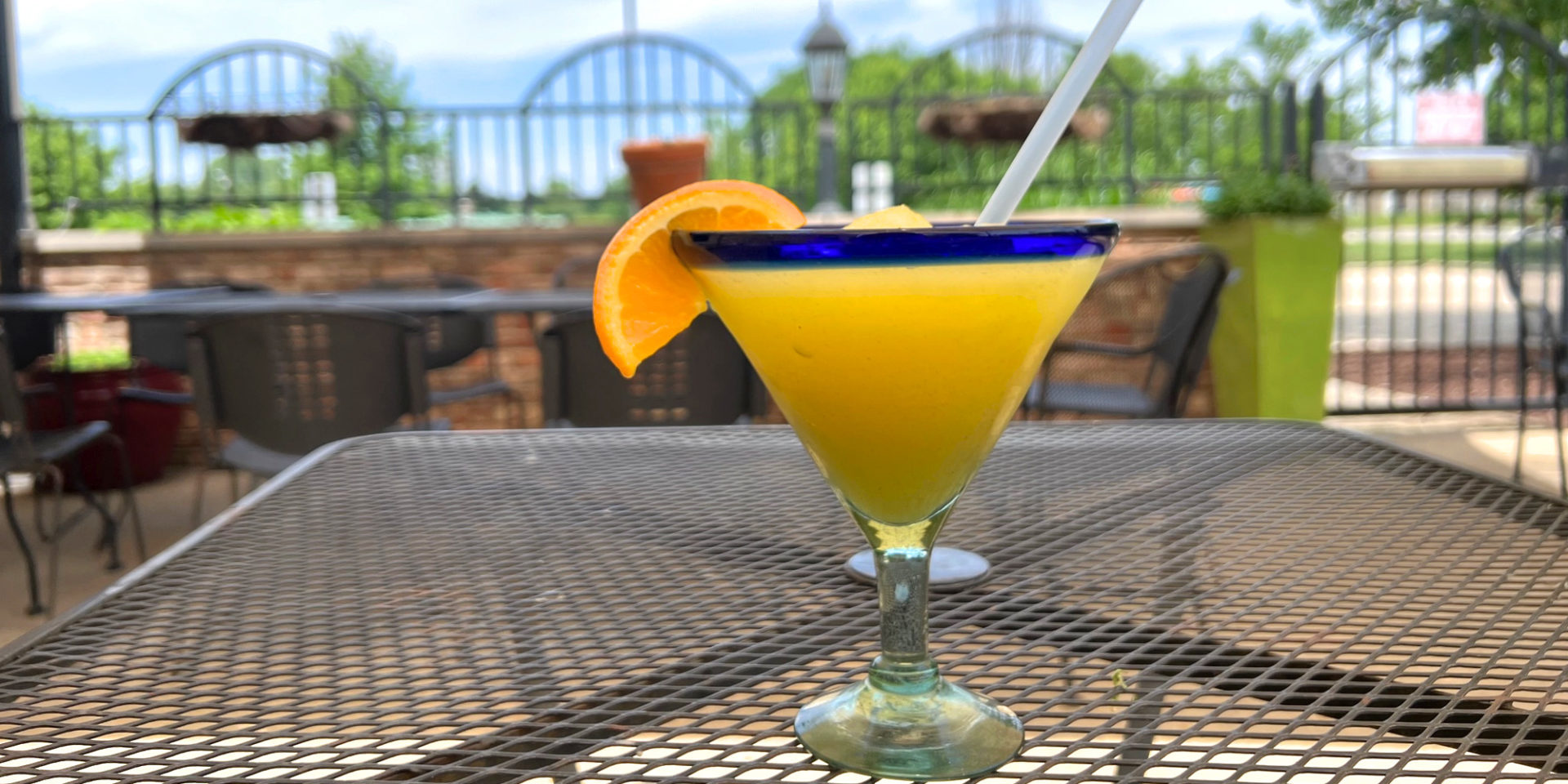 On a patio, there is a pineapple margarita with an orange slice in a martini shaped margarita glass with a blue rim. Photo by Alyssa Buckley