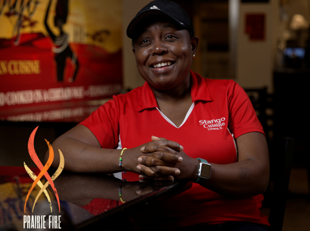 A Black woman wearing a black cap and red shrit is leaning against a dark countertop and smiling.