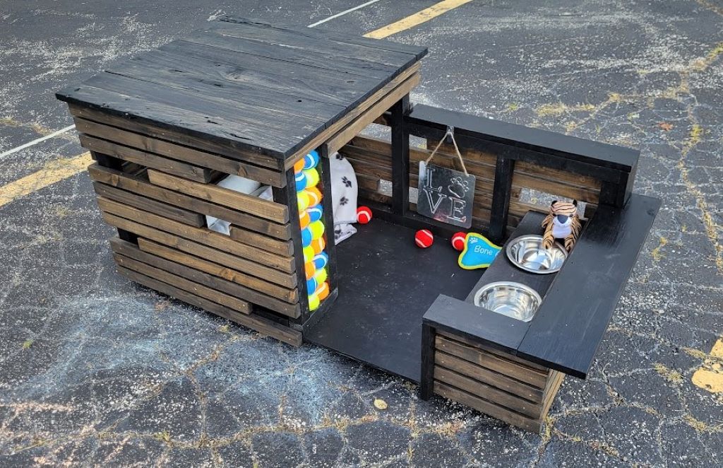 A dog house with wooden slats along the side and a flat gray roof. There is a terrace area in front of the opening that has various toys scattered around it.