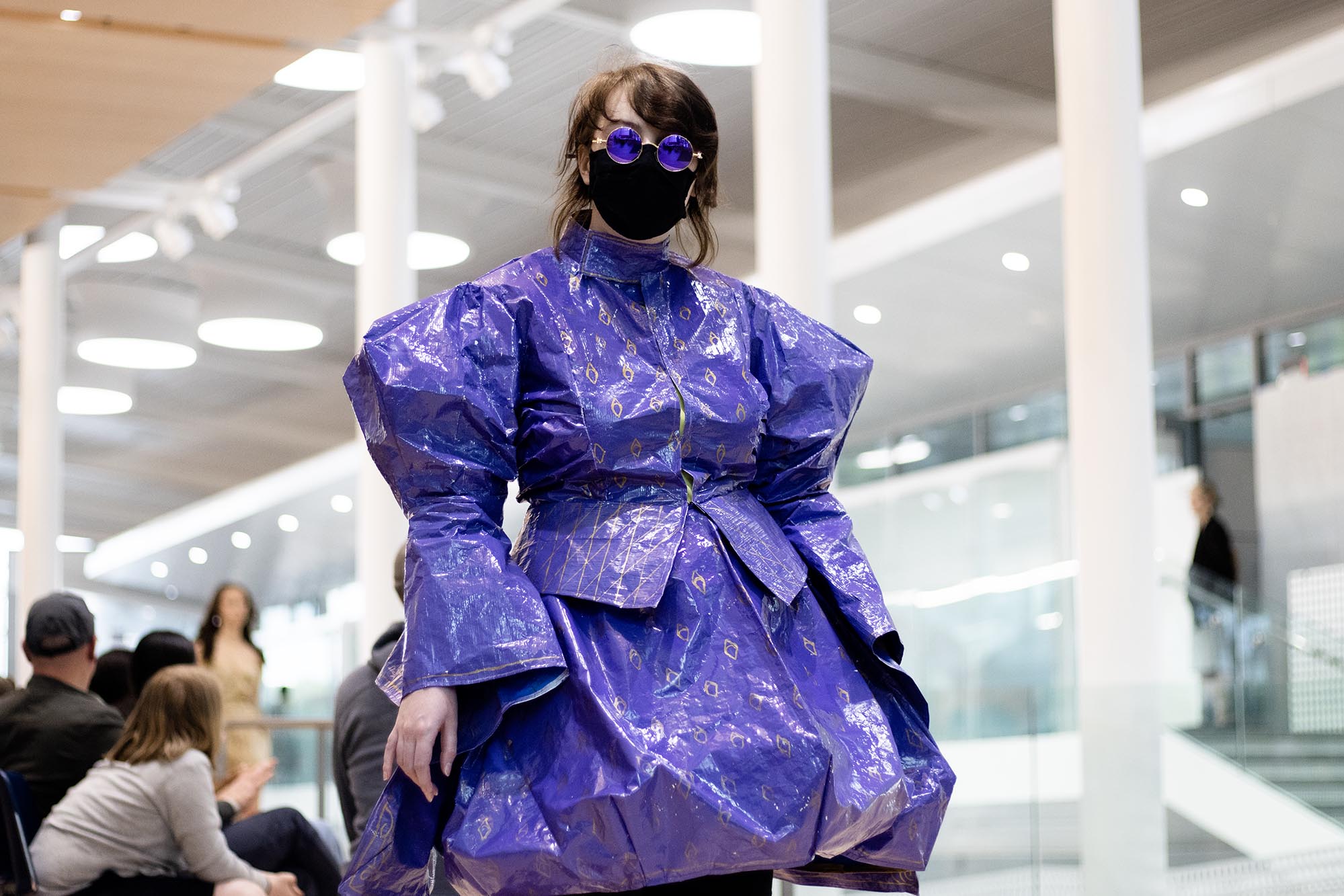 A model wears a purple suit jacket and skirt made from tarp or vinyl billboard. The female model also wears a black face mask and round purple glasses.