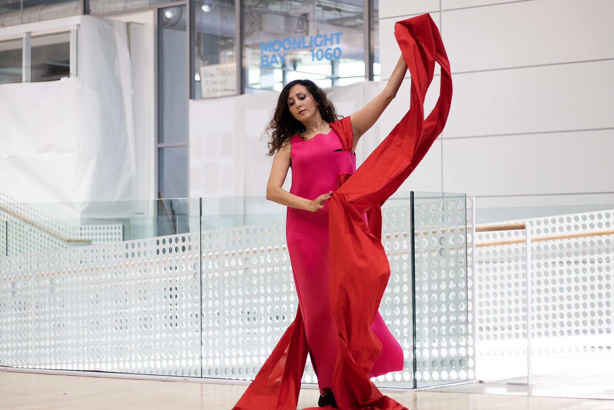 Re-Fashioned 2023. Model Banafsheh wears a long, hot pink dress with a red fabric train that comes over her shoulder. Her left arm holds the red fabric up in the air. Designed by Natalia Espinel-Porras.