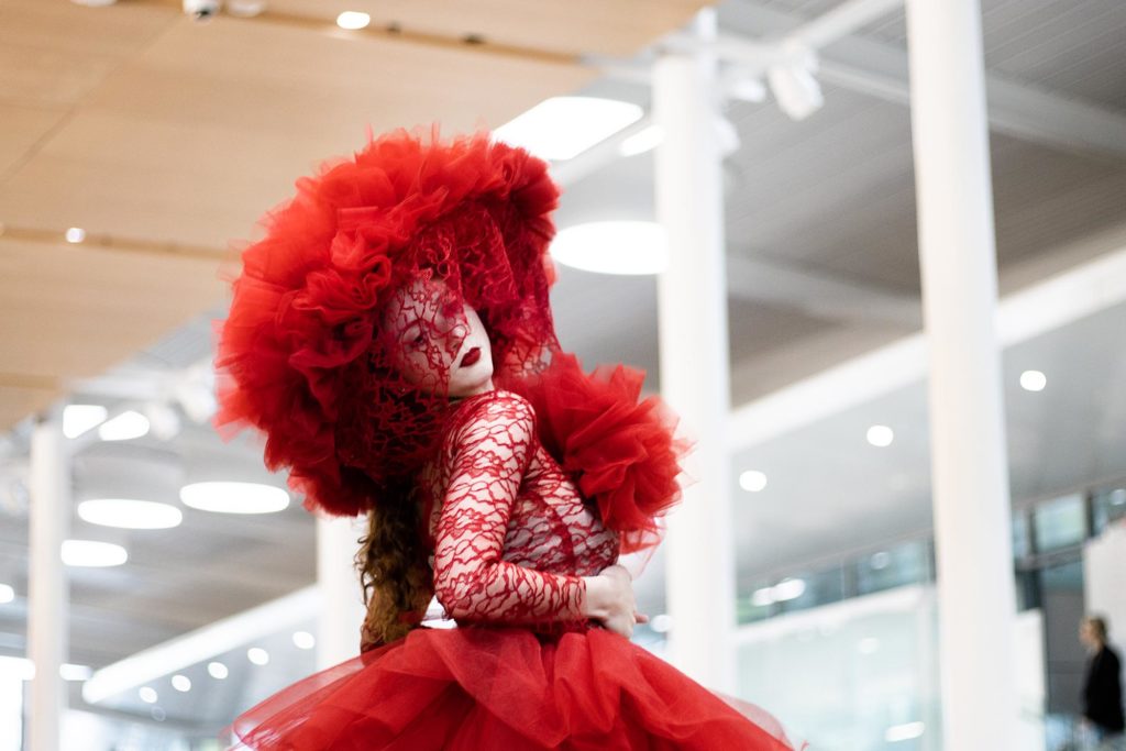 Re-Fashioned 2023. Model Micheala Burrows wears a red outfit with a huge tulle head piece and lace veil over the face. The top of the outfit it the same red lace over white fabric, with a large red tulle skirt. Designed by Miranda Mottlowitz.
