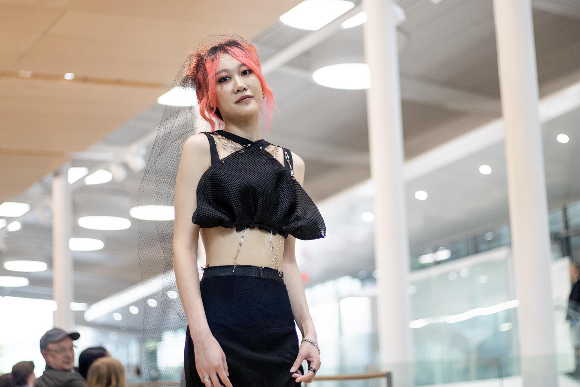 Re-Fashioned 2023. Model Vicky Ma wears a black cropped top and black bottom with metal adornments. She has pink hair and a black lace veil. Designers Alan Dong and Yi Liu.