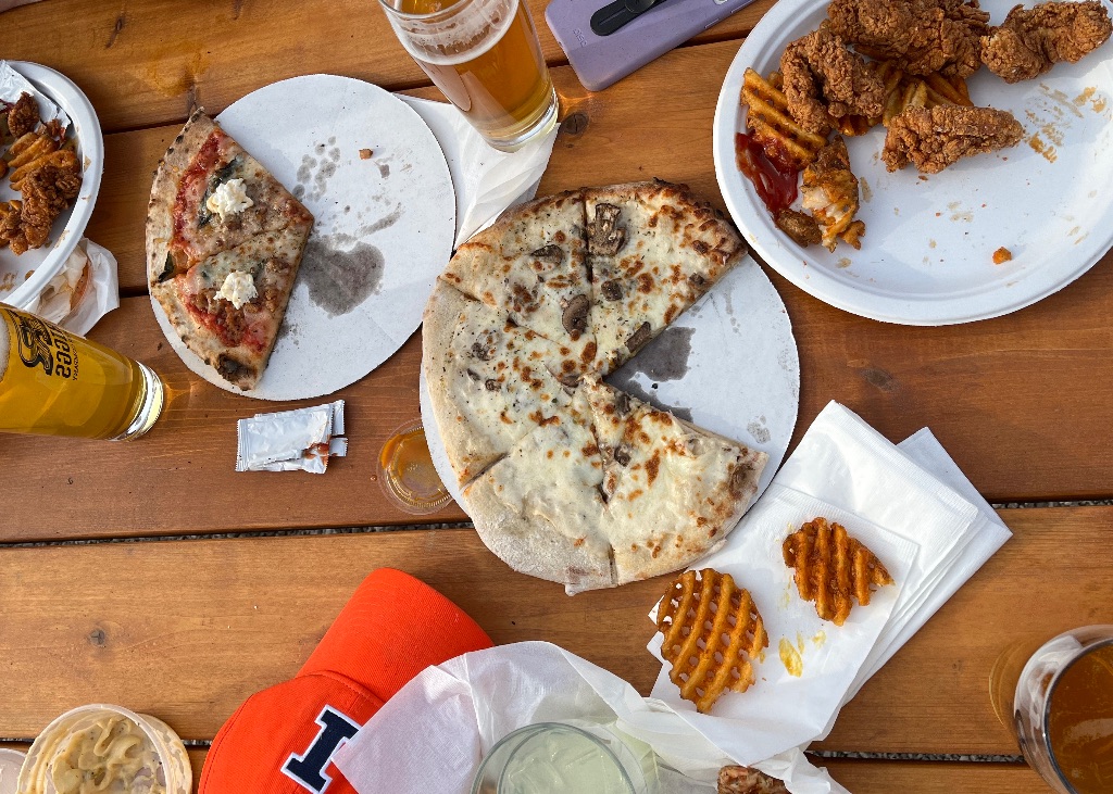 A picture from above a wooden table with plates of pizza, chicken tenders, and waffle French fries. The table also has big pints of beer, an orange baseball hat with a blue I on it, napkins, and a purple phone.