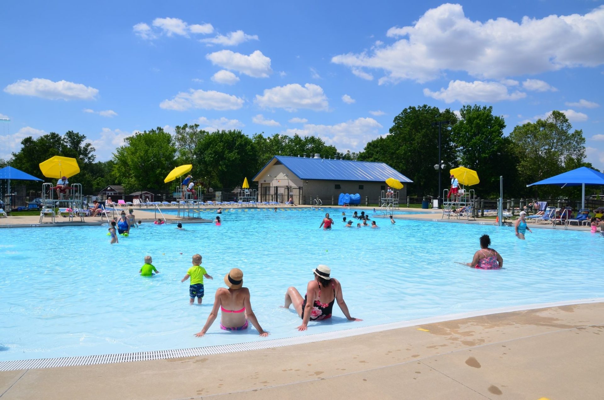 Wide shot of a large swimming pool with zero entry. There are people scattered throughout, and lifeguard chairs with yellow umbrellas at various spots around the pool.