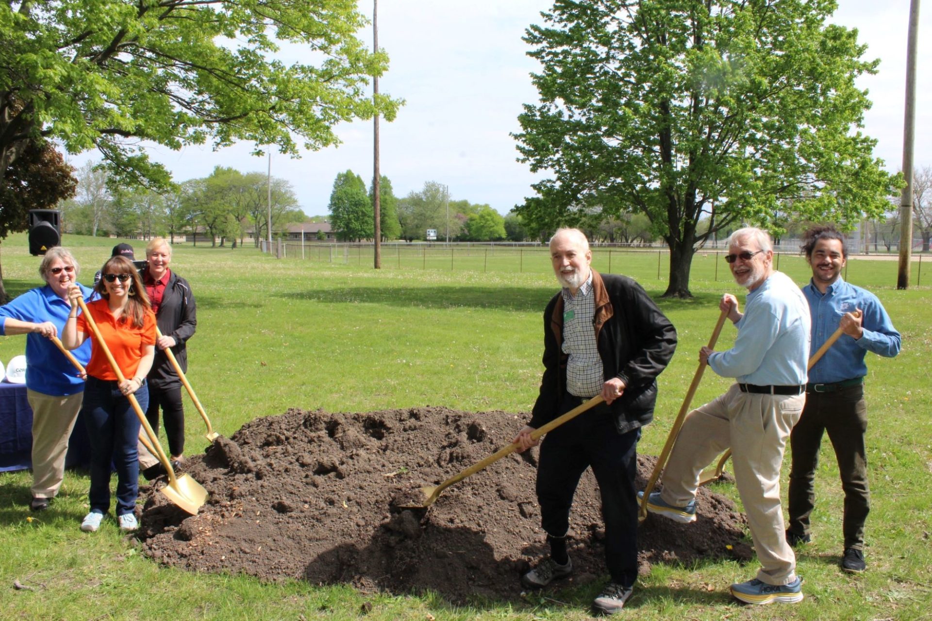 Six people are gathered around a pile of dirt, in the middle of a large grassy area. They are each holding a shovel.