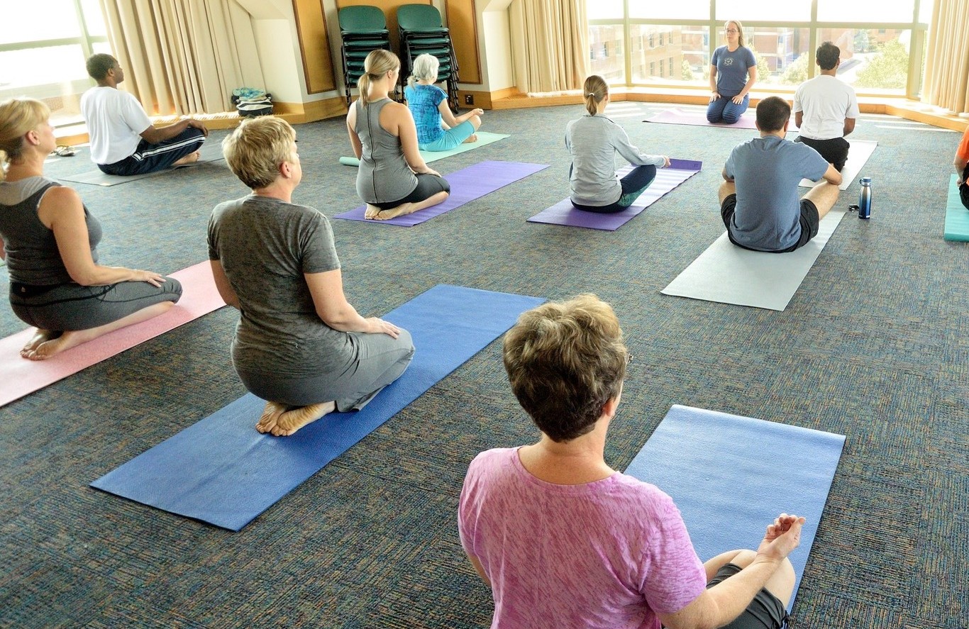 There are several people kneeling on yoga mats, spread out throughout a large room with windows on either side.