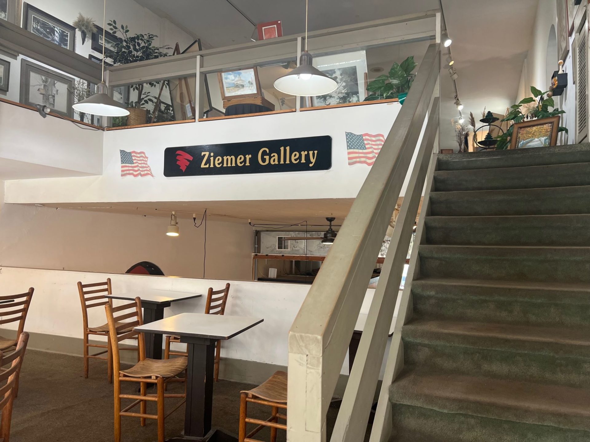 A set of gray carpeted stairs lead up to an art gallery. On a white wall below the gallery there is a black sign that says Ziemer Gallery in gold lettering.