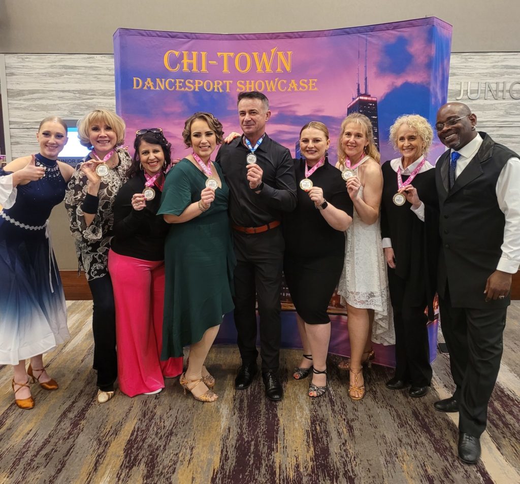 Cinnamon Trammell, a Black man in dress pants, a white shirt and vest, stands alongside students after a dance showcase. A banner behind them reads "Chi Town Dancesport Showcase." The students are holding up medals. 