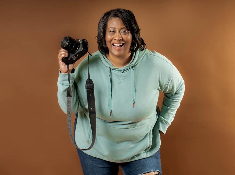 A black woman with dark shoulder length hair wears a pale green sweatshirt and ripped jeans. She stands against a brown backdrop and is holding a digital camera..