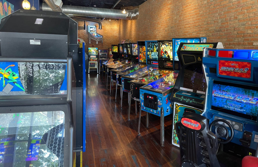 View of narrow room with brick walls. On the right are a bunch of pinball machines and arcade games. On the left are larger, driving-style arcade games.