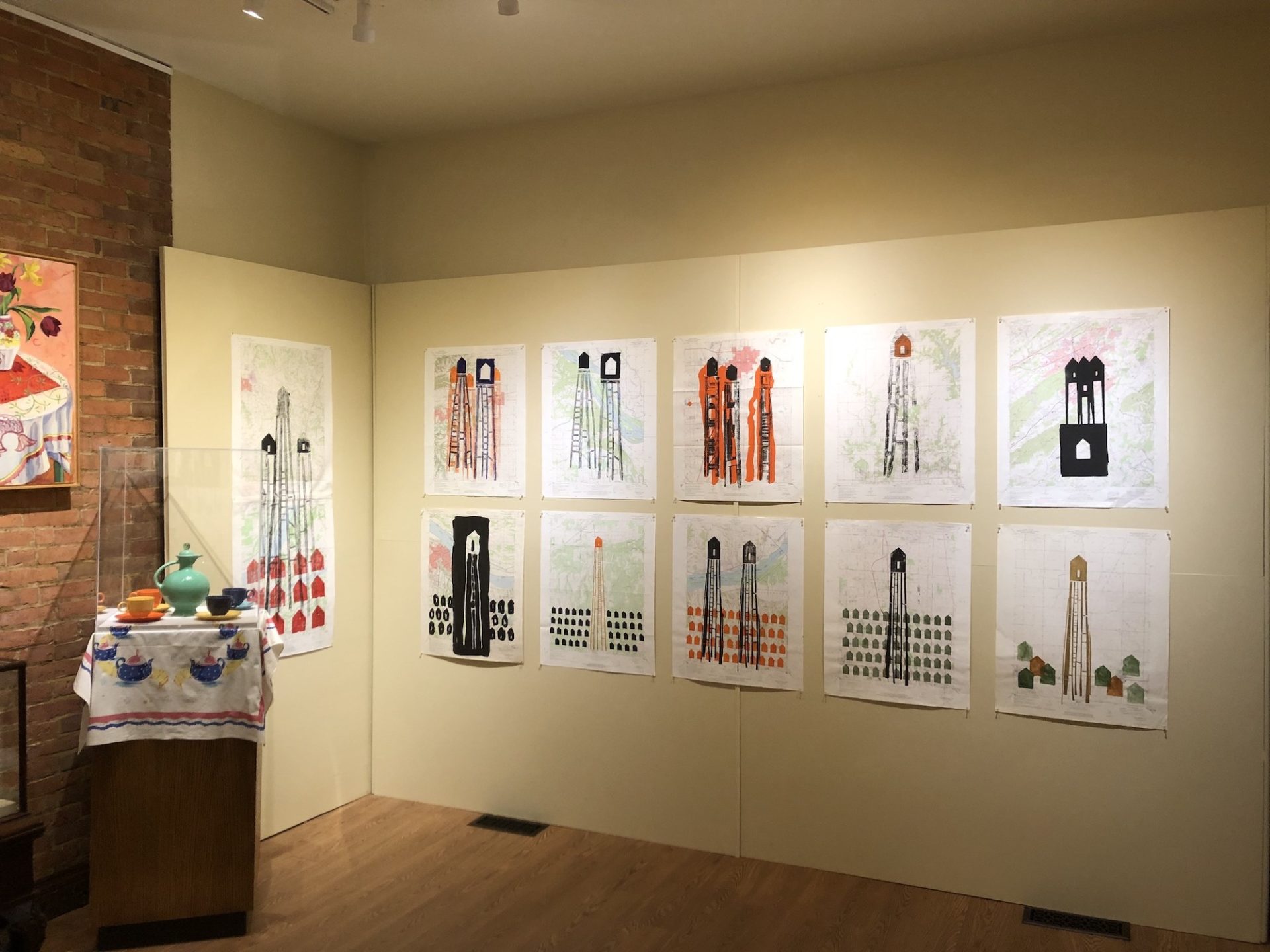 10 of Robb Springfield's Stilt Houses creations are on a gallery wall in 2 rows of 5 works each. The stilt house images are large and bold in black, red, orange, and blue. The background of each picture is an Illinois map. 