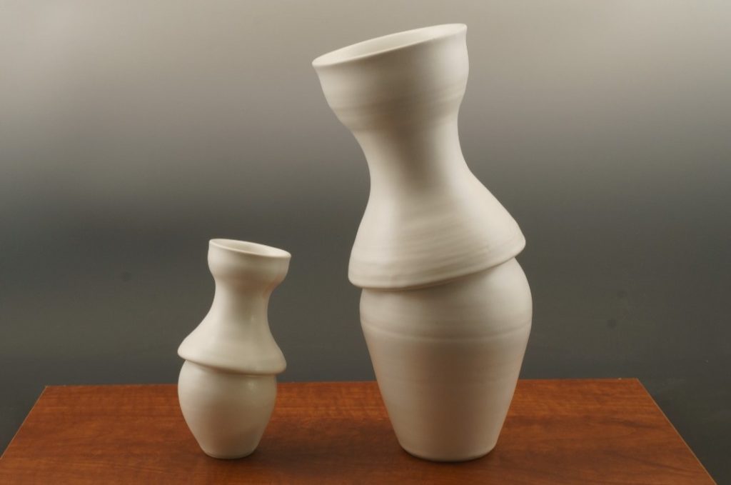 2 white vases, each has two sections. the one on the right is a little less than twice the height of the one on the left, the vases have the appearance of leaning towards each other through their top halves. 