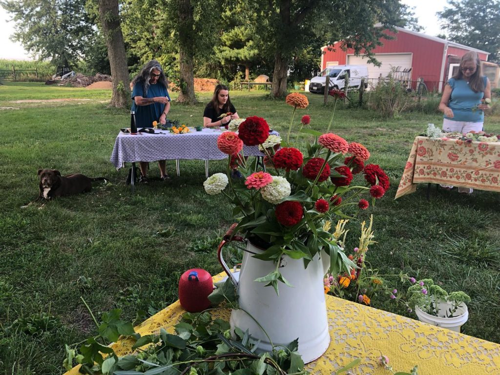 A flower arranging workshop outdoors. In the foreground, a white metal pitcher with red, pink, and white flowers in it sits on a table with a yellow lacy tablecloth. In the background are tables with people working to arrange flowers. There is a red barn in the background.