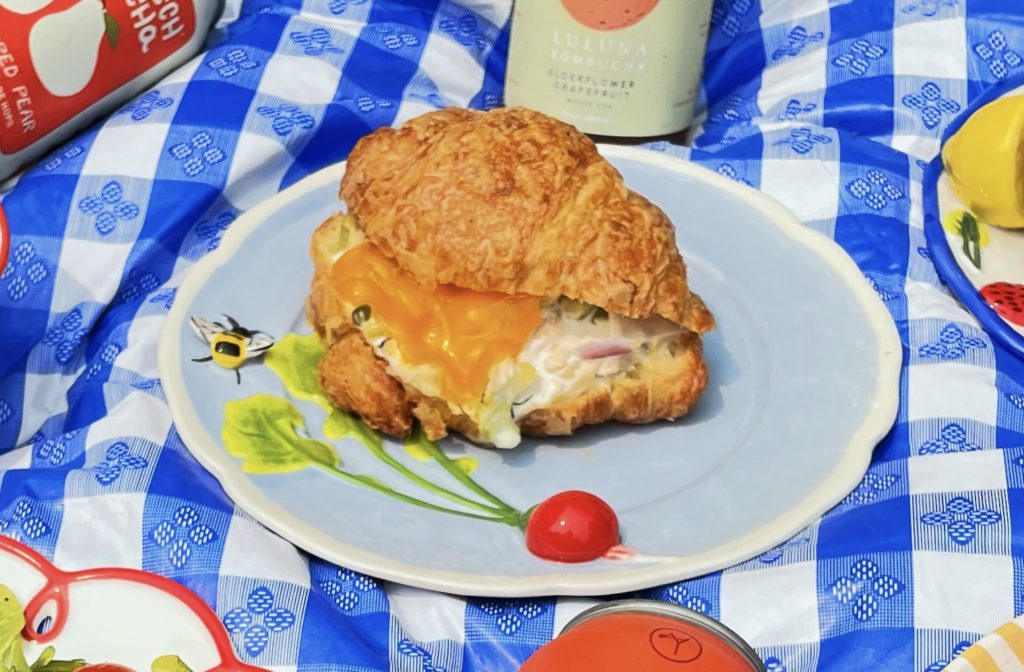 Hopscotch: A croissant sandwich sits on a blue plate. Tuna salad and melted cheddar cheese visible inside the croissant bun. The plate sits on a blue gingham tablecloth and there are various canned beverages peeking into frame.