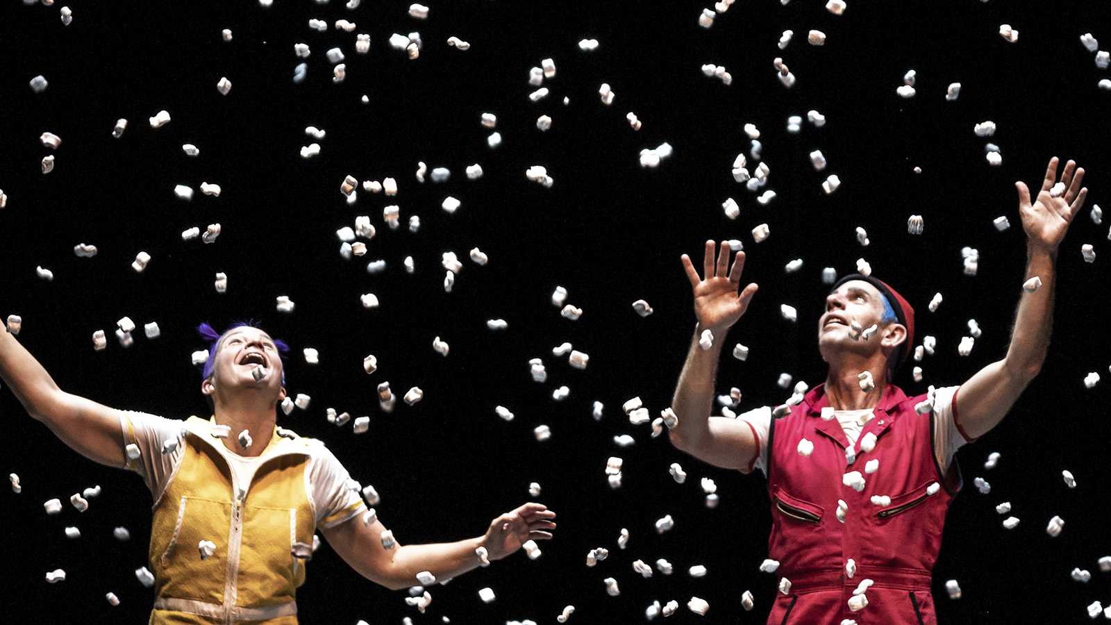 Acrobuffos; two people stand on stage. The person on the left has purple hair and is wearing a yellow zip up jump suit, the person on the right has blue hair and is wearing a matching red jumpsuit. What appears to be packing popcorn is falling from the ceiling. The background is entirely black