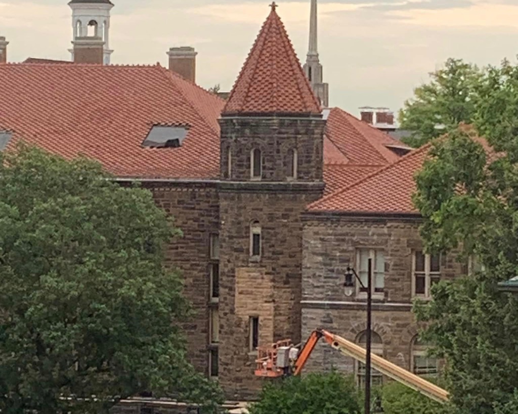 A brown brick building with a circular turret and a red roof is under construction. Some of the stones have been replaced with new and lighter stone. There are trees on either side of the image and a construction crane in the bottom right corner.