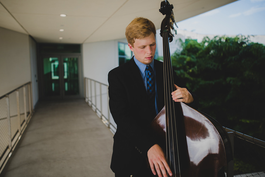 A man dressed in a suit holding an upright bass and looking down in a hallway